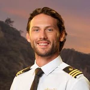 Alex Wright wearing his Below Deck uniform and standing in front of a sunset